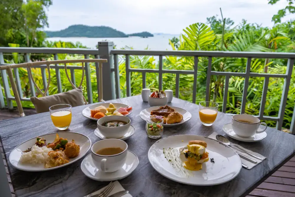 A beautiful breakfast spread for two, including eggs benedict and fresh fruit, set on a balcony table overlooking a lush tropical landscape and the sea, representing a unique Seminyak cafe experience.