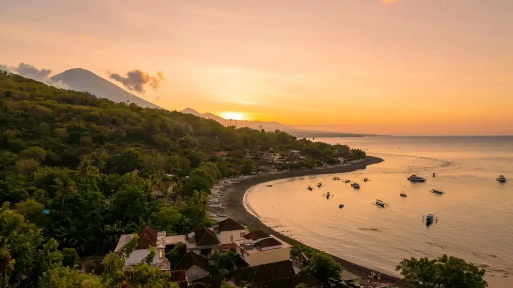 A golden sunrise over the quiet northeast coast of Bali, showing Amed's curving black sand beach, traditional jukung fishing boats in the bay, and the majestic Mount Agung volcano in the background.