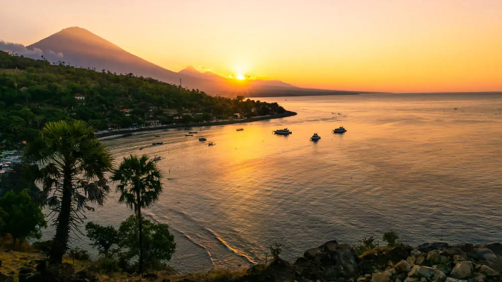 A stunning sunrise over the Amed coastline in Bali, with the golden sun rising behind the majestic Mount Agung volcano and casting its light across the calm sea and fishing boats.