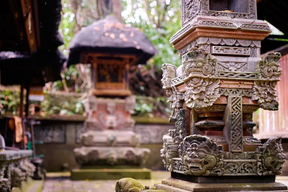 A close-up of an ornate, moss-covered stone carving at a Balinese temple, a key element of traditional Balinese architecture. The background shows another traditional shrine in a lush setting.