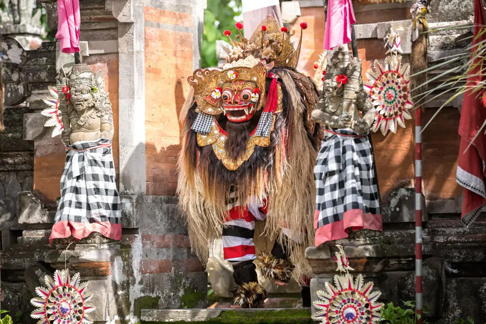 A performer in a traditional Barong costume, featuring an ornate mask and hairy body, during a Balinese dance performance in a temple gateway.