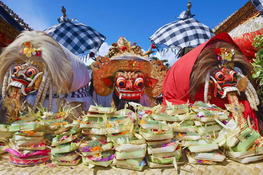 Sacred masks of Barong and Rangda at a temple ceremony, central to Balinese Hinduism. In front of the masks are dozens of canang sari, or daily offerings, with traditional Balinese umbrellas in the background.