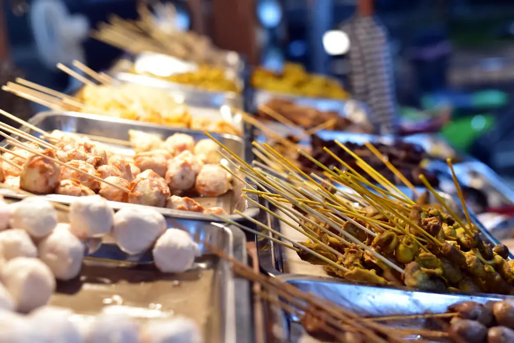 A close-up of a street food stall at a Bali night market, with metal trays filled with various types of sate skewers, including fish balls and chicken satay.