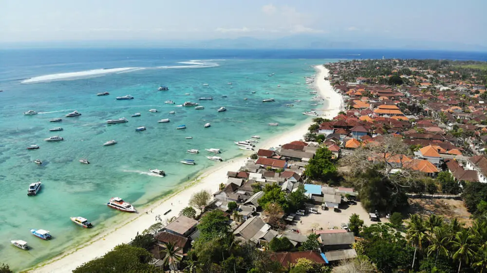 An aerial view of Jungut Batu beach on Nusa Lembongan, Bali, showing the long white sand beach, the village, and dozens of boats moored in the shallow turquoise water.