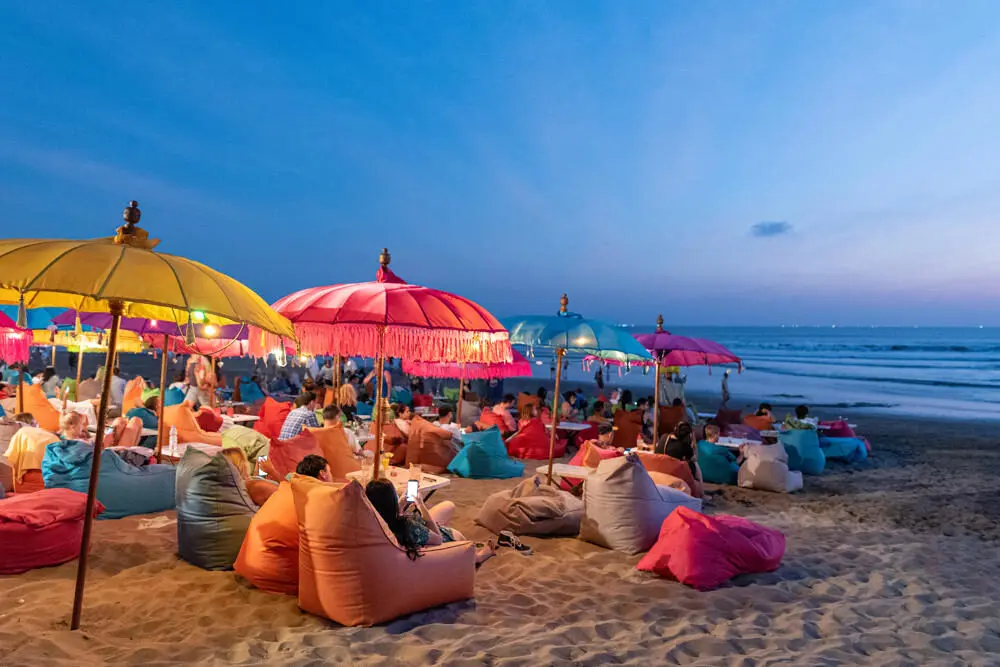 People relaxing on colorful beanbag chairs at a beach club in Seminyak, Bali, watching the ocean at dusk under vibrant umbrellas.