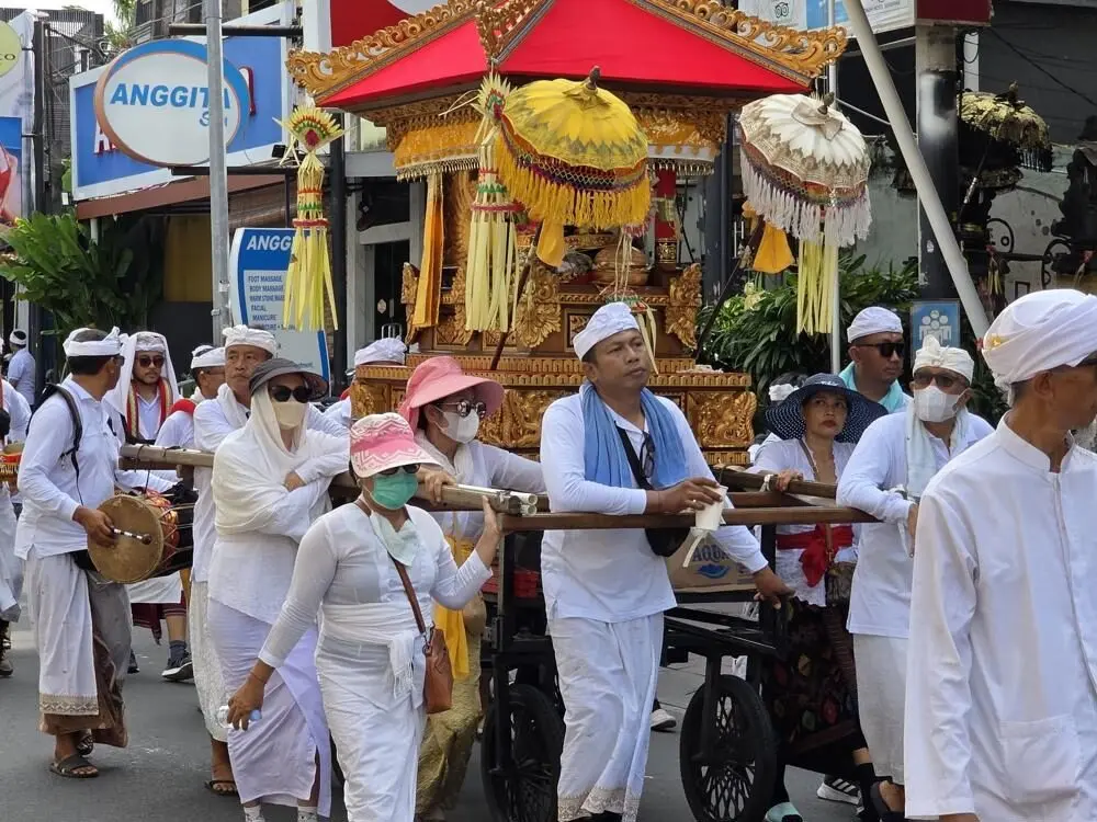 Balinese people in traditional white dress participating in a Melasti procession, one of the island's most important festivals and ceremonies, carrying an ornate golden shrine down a street.