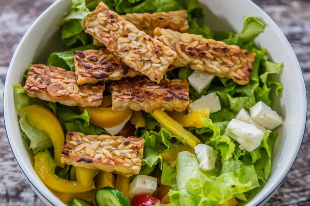 A close-up view of a vegan tempeh goreng salad in a white bowl, a popular plant-based dish in Bali. It features crispy fried tempeh on a bed of fresh lettuce with bell peppers and vegan cheese.