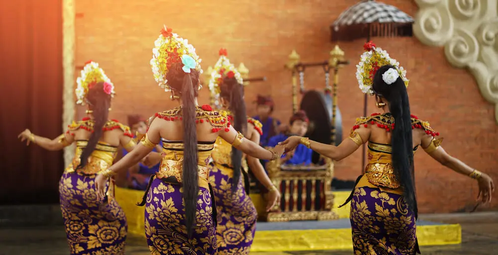A group of Balinese dancers seen from behind, showcasing their intricate purple and gold costumes and golden headdresses during an authentic cultural performance in Bali.