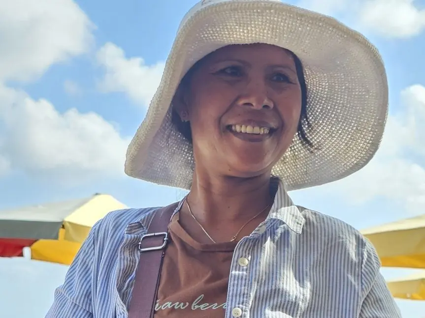 A close-up portrait of Angela Cindy, a Balinese beach seller, smiling warmly in the sun while wearing a wide-brimmed white hat on Legian beach.