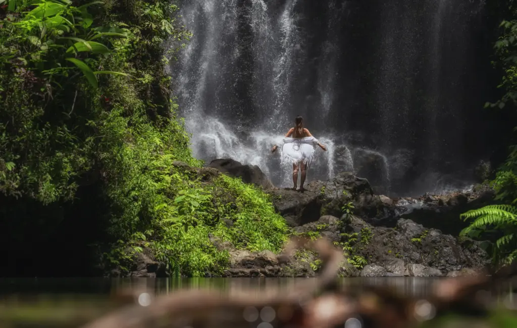 A woman in a white dress stands on a rock with her arms out, facing a powerful waterfall in a lush, green jungle setting.