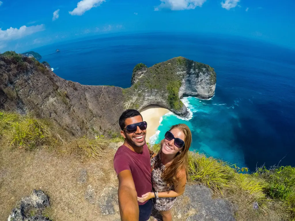 A happy couple wearing sunglasses taking a selfie on a clifftop with the iconic T-Rex shaped cliff of Kelingking Beach and the bright blue ocean in Nusa Penida in the background.
