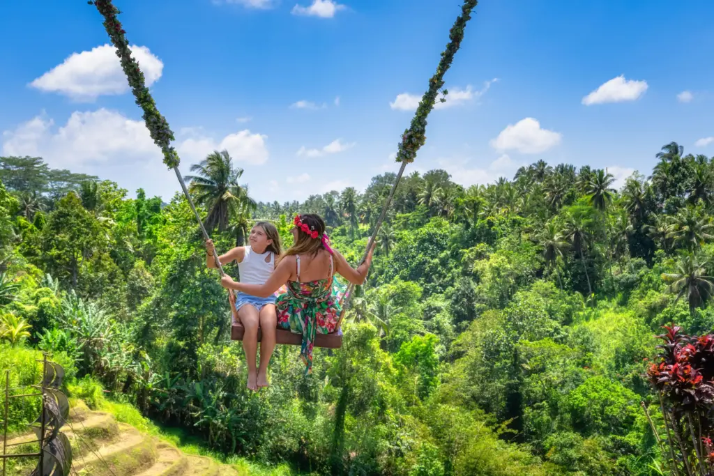 A mother and her daughter are sitting together on a large swing, soaring high above the lush green jungle landscape in Bali, both smiling and enjoying the ride.
