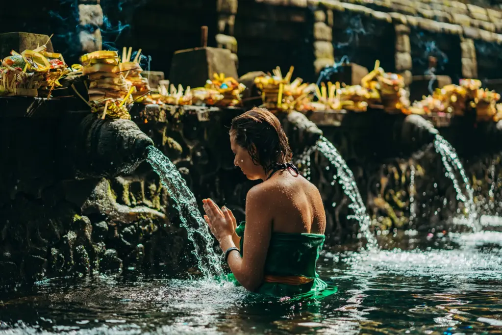 "A woman in a green sarong, with her hands pressed together in prayer, performs a water purification ritual under a stream of water at a Balinese temple. "