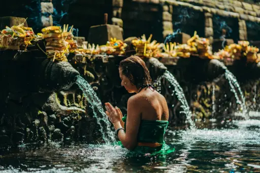 "A woman in a green sarong, with her hands pressed together in prayer, performs a water purification ritual under a stream of water at a Balinese temple. "