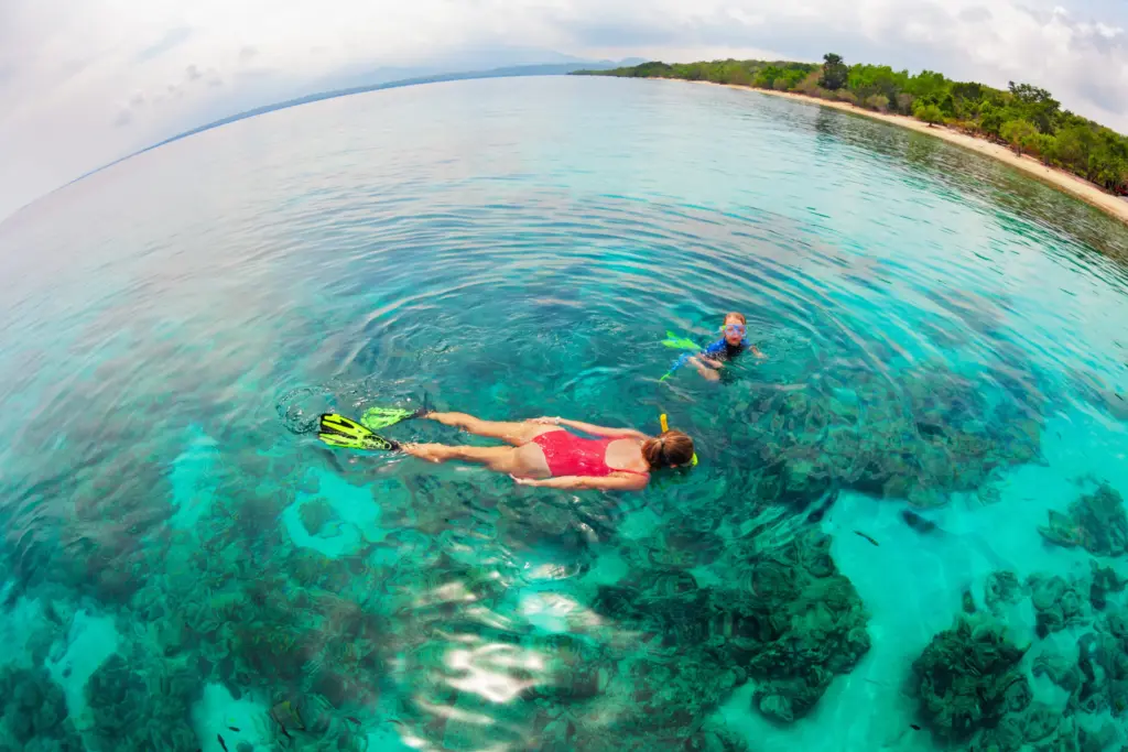 A mother and child are snorkeling in clear, turquoise water over a coral reef near a tropical island beach