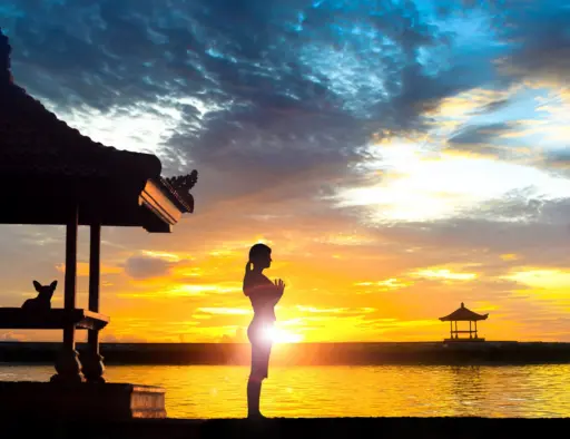 A silhouette of a woman standing in a yoga pose with her hands together at her chest, facing a beautiful golden sunset over the ocean on a beach in Bali.