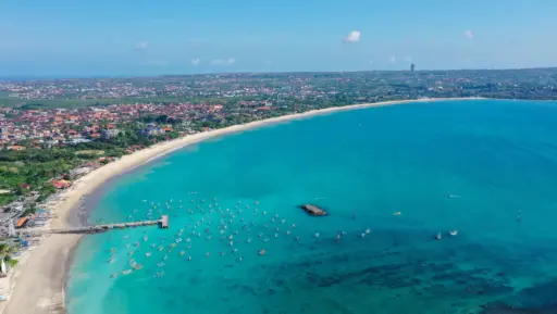 An aerial shot of Jimbaran Bay showing a long, sandy beach, calm turquoise water, a pier, and many traditional fishing boats (jukung) anchored near the shore.