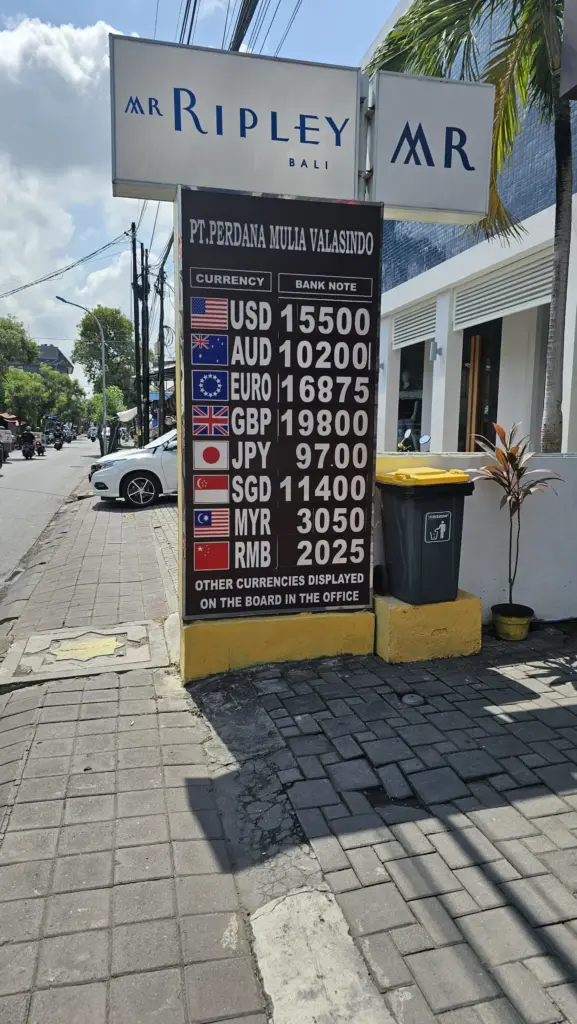 A street-level photo of a reputable money changer in Bali with a clear sign displaying currency exchange rates for USD, AUD, EUR, GBP, SGD, MYR, and RMB, next to a building with a "Mr Ripley" sign.