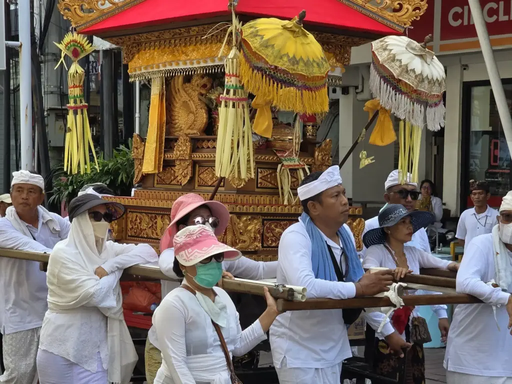 A group of Balinese people in traditional white clothing carry a gilded, canopied structure during a religious procession in a street.