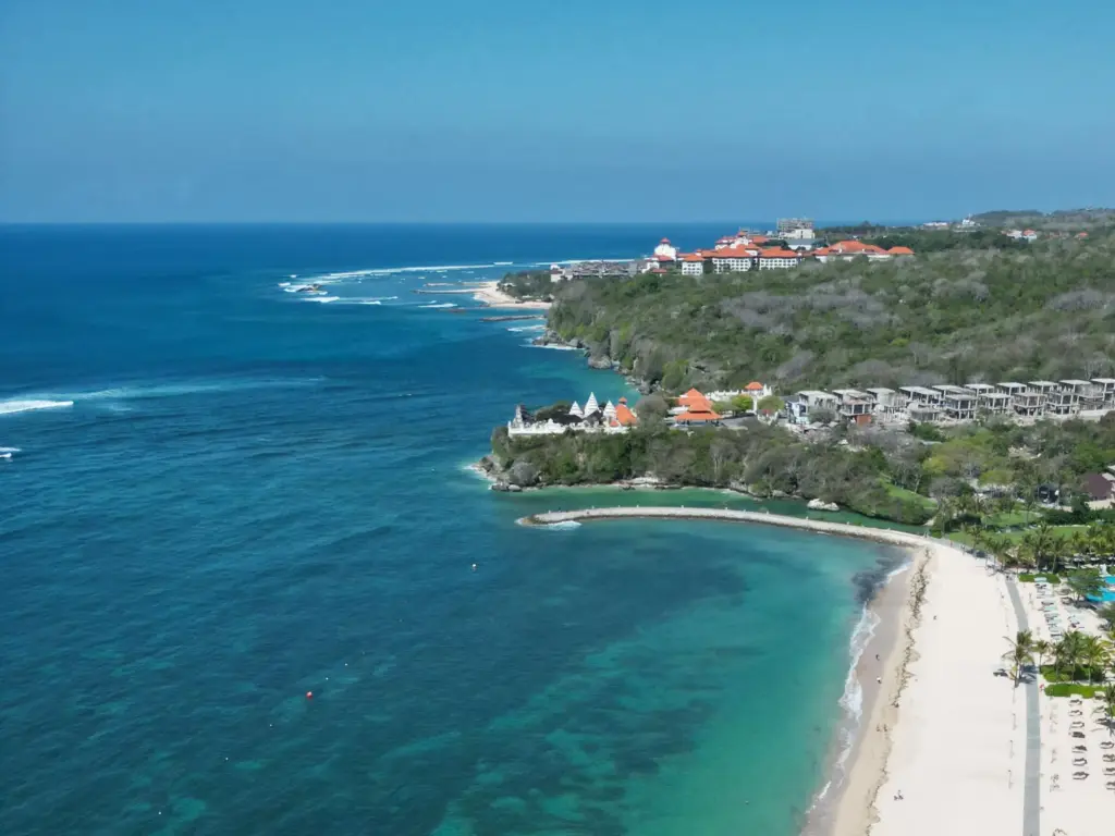 An aerial view of Geger Beach in Nusa Dua, showing a calm, clear turquoise bay, a long stretch of white sand, and beachfront resorts surrounded by lush green trees.