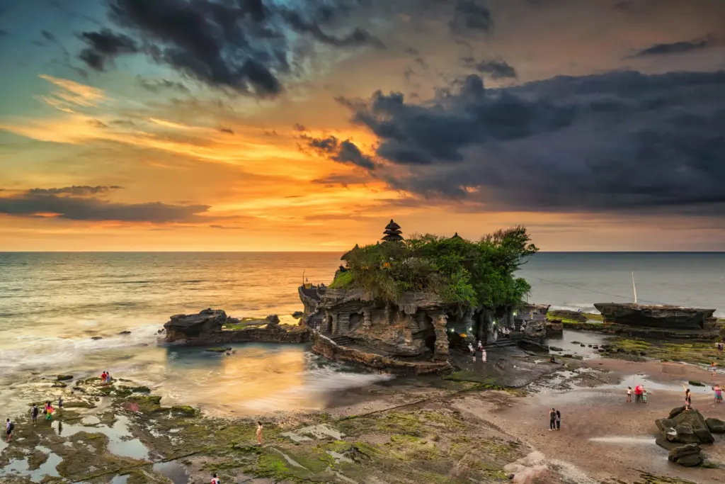 A dramatic sunset over Tanah Lot Temple, with the iconic temple on a rocky outcrop surrounded by water and people visible on the beach in the foreground.