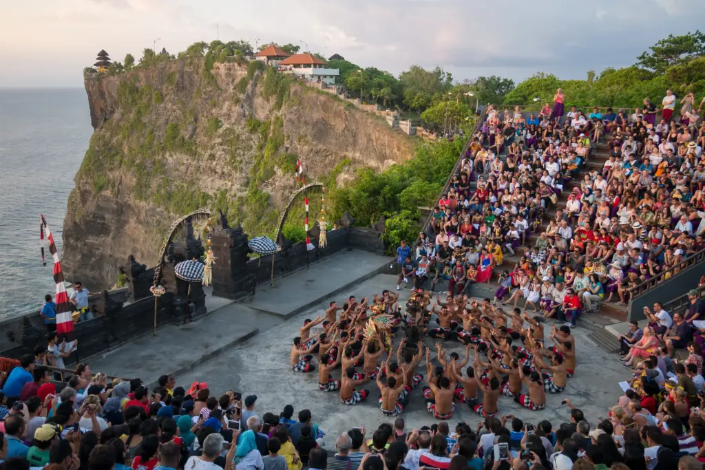 A wide-angle view of a traditional Balinese Kecak dance performance with dozens of male dancers chanting in a circle on a stage at Uluwatu Temple, with a crowd of spectators and a cliff overlooking the ocean at sunset.