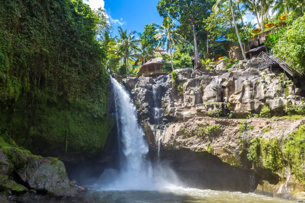 A wide-angle view of the Tegenungan Waterfall cascading into a pool surrounded by lush green jungle foliage, palm trees, and rocks under a bright blue sky.