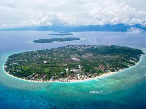 An aerial view of Gili Trawangan showing its white-sand beaches, clear turquoise water, and a densely vegetated interior, with the smaller Gili Meno and Gili Air visible in the background.