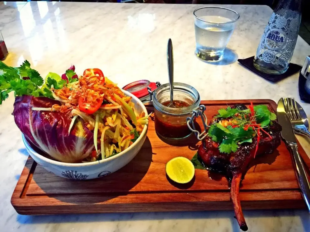 A close-up of a beautifully plated Balinese meal on a wooden board, featuring a glazed pork chop and a side salad.