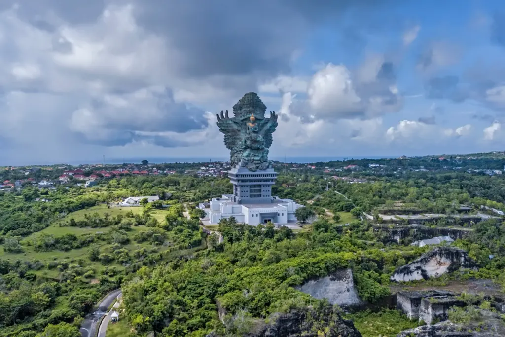 An aerial shot of the massive Garuda Wisnu Kencana statue on a pedestal, surrounded by lush green hills, with the coastline and ocean visible in the background under a cloudy sky.