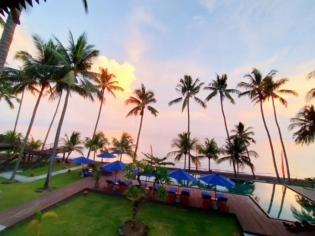 Oceanfront view of Kirana Tembok in Bali with palm trees, pool, and sunset sky