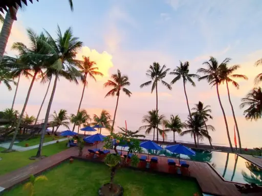 Oceanfront view of Kirana Tembok in Bali with palm trees, pool, and sunset sky