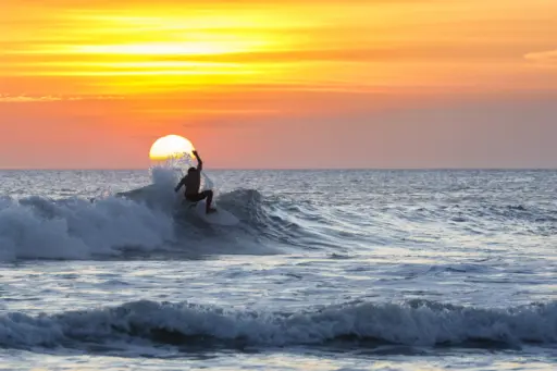 A silhouette of a person on a surfboard, riding a wave as the sun sets over the ocean in Kuta, Bali.