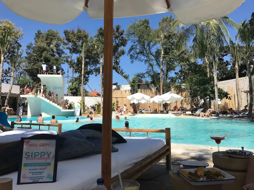 A view of a lively beach club with a large swimming pool, a high diving board, and people lounging on daybeds under umbrellas.