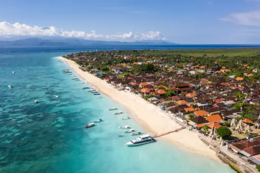 An aerial view of Jungut Batu, Nusa Lembongan, showing the long white-sand beach, boats docked in the clear turquoise water, and the coastal village nestled among palm trees.
