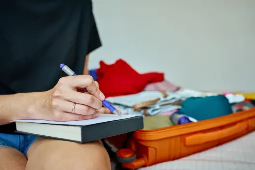 A close-up of a person's hands writing a checklist in a notebook, with an open suitcase full of clothes and a pair of shorts in the background, representing trip planning.