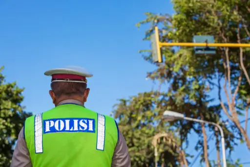 A back-view of a uniformed Indonesian police officer ("POLISI") in a neon green vest, standing in the middle of a street in Bali with trees and a blue sky in the background