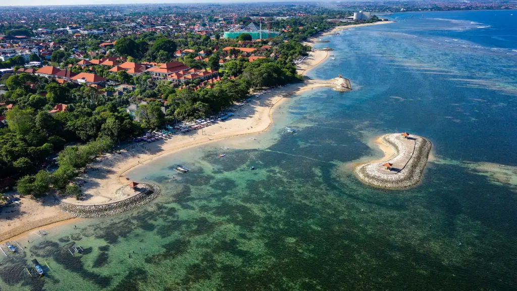 An aerial shot of the calm, clear turquoise waters and soft golden sand of Sanur Beach, showing small breakwaters and resorts nestled among green trees.