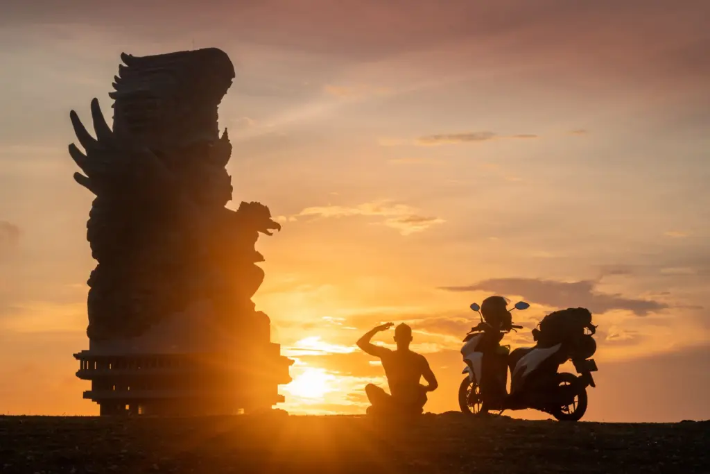 A silhouetted image of a person sitting cross-legged next to a scooter, saluting a massive stone statue against a vibrant orange and yellow sunset sky in Bali.