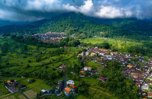 An aerial shot of the Sidemen valley, showing a traditional village with red-tiled roofs nestled amongst lush green rice fields and hills, with clouds hanging over the mountains.