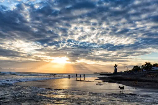 A beautiful sunset over Pererenan Beach, with sun rays breaking through a cloudy sky and silhouettes of people and dogs on the wet sand at the water's edge.