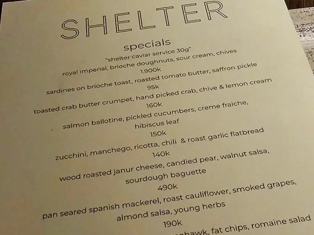 A close-up photo of a menu at a Bali restaurant called "Shelter", with prices for specials like "royal imperial caviar" and "toasted crab butter crumpet", illustrating how costs are listed before tax and service fees.