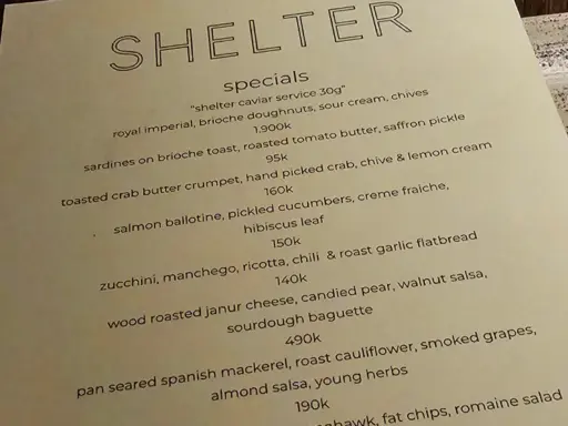 A close-up photo of a menu at a Bali restaurant called "Shelter", with prices for specials like "royal imperial caviar" and "toasted crab butter crumpet", illustrating how costs are listed before tax and service fees.