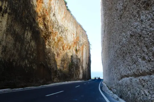 A dramatic road carved into tall, textured limestone cliffs on the way to the beach in Uluwatu, with a view of the ocean at the end.